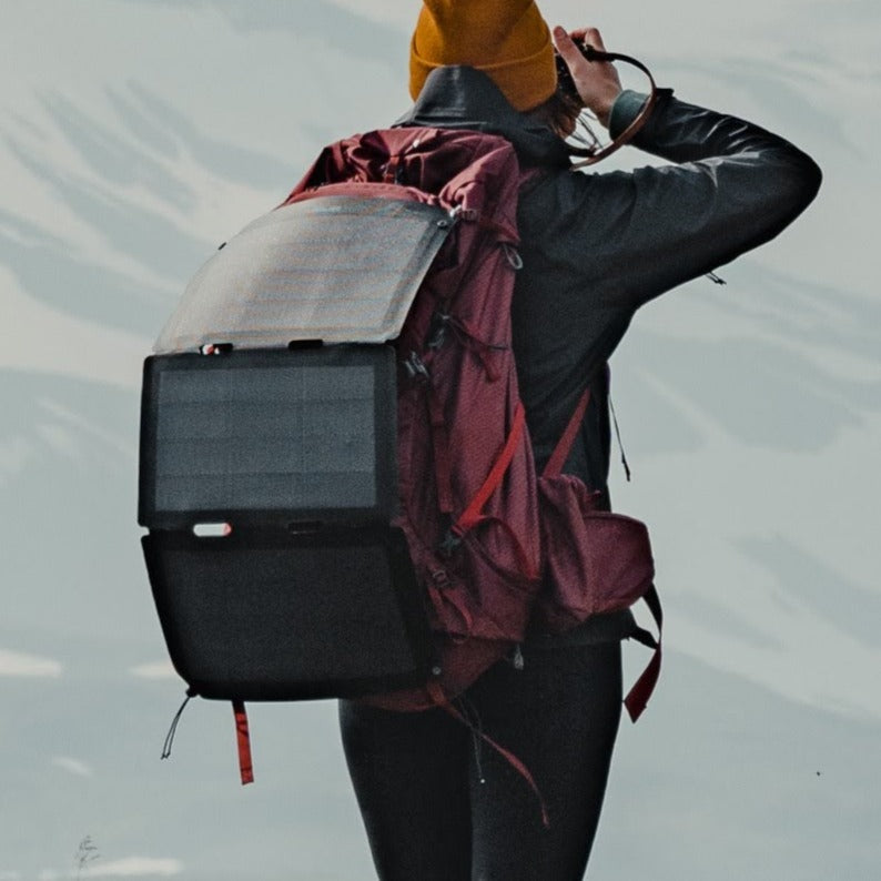Frau auf dem Gipfel eines verschneiten Berges mit Kamera in der Hand und faltbarem Solarpanel, das an ihrem Rucksack befestigt ist