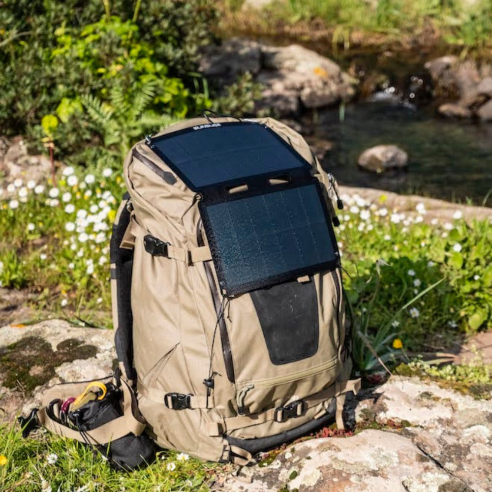 sac de randonnée posé au bord d'un ruisseau, auquel est attaché un panneau solaire portable pour le camping