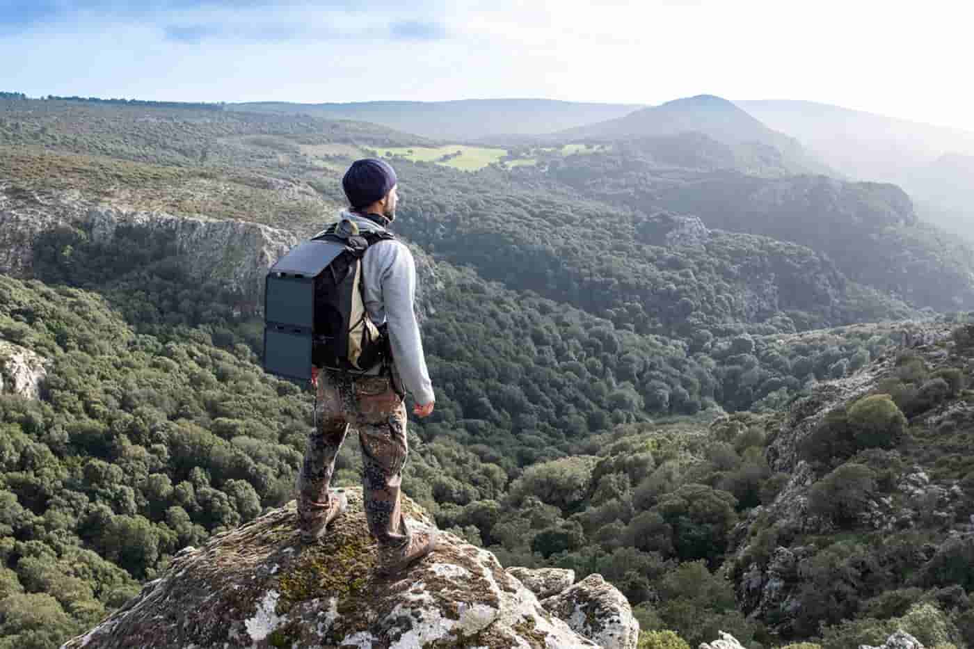 ein Mann auf dem Gipfel eines sonnigen Berges mit einem an seinem Rucksack befestigten Wandersolarpanel