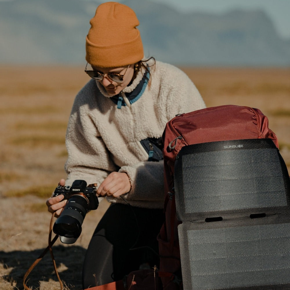 Frau, die ein Foto mit einem an ihrem Rucksack befestigten Wandersolarmodul auf dem Boden machen will