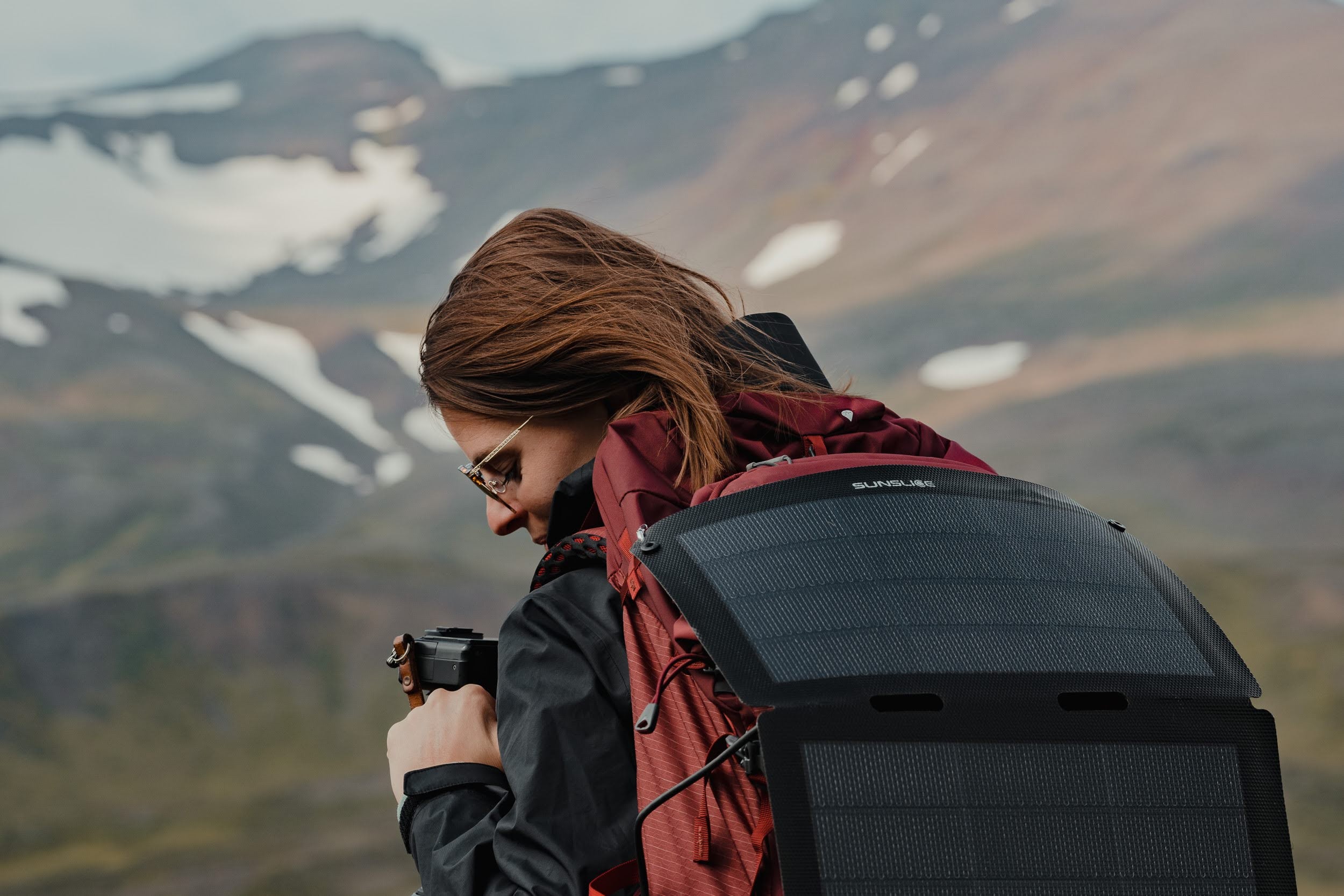 vrouw op de top van een besneeuwde berg met camera in de hand en opvouwbaar zonnepaneel bevestigd aan haar rugzak