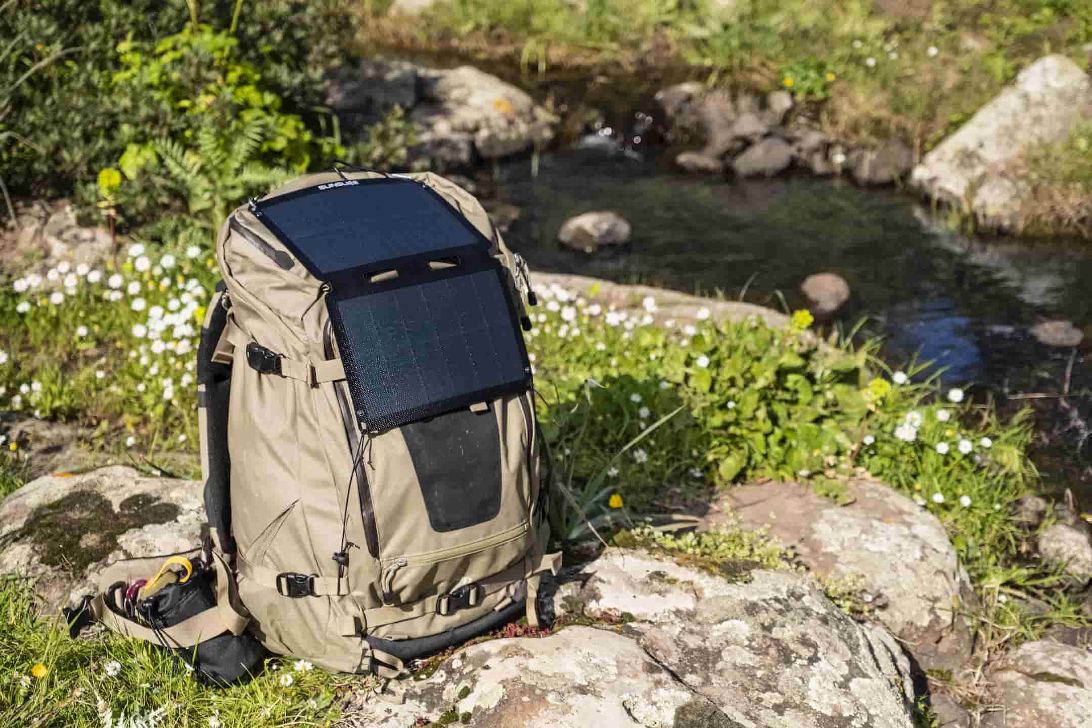 sac de randonnée posé au bord d'un ruisseau, auquel est attaché un panneau solaire portable pour le camping