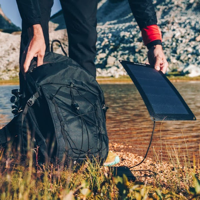 Mann mit tragbarem Solarmodul für das Camping, das er an seinem Rucksack befestigt hat, auf einem Berg mit grünem Hintergrund am Horizont