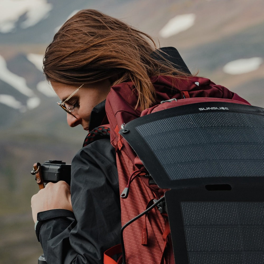 Frau auf dem Gipfel eines verschneiten Berges mit Kamera in der Hand und faltbarem Solarpanel, das an ihrem Rucksack befestigt ist